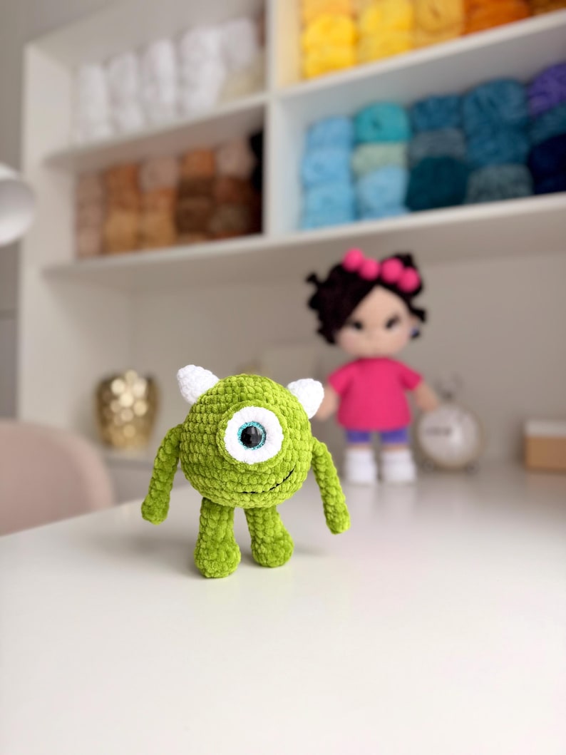May include: A small, round, green crocheted monster plush toy with a single large central eye, two small white horns on top, and two thin arms and legs. The creature has a textured yarn surface and a stitched smile on its round body.
