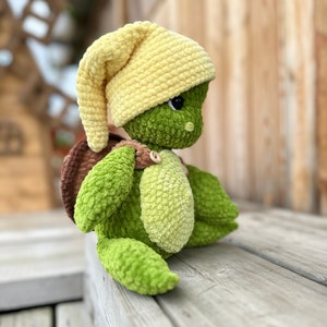 Puede incluir: Un juguete de peluche con forma de tortuga hecho de hilo verde y verde claro, con un caparaz&oacute;n marr&oacute;n y un gorro amarillo suave. La tortuga tiene una cabeza redonda con ojos negros, una mancha verde claro en el vientre y un bot&oacute;n de madera.