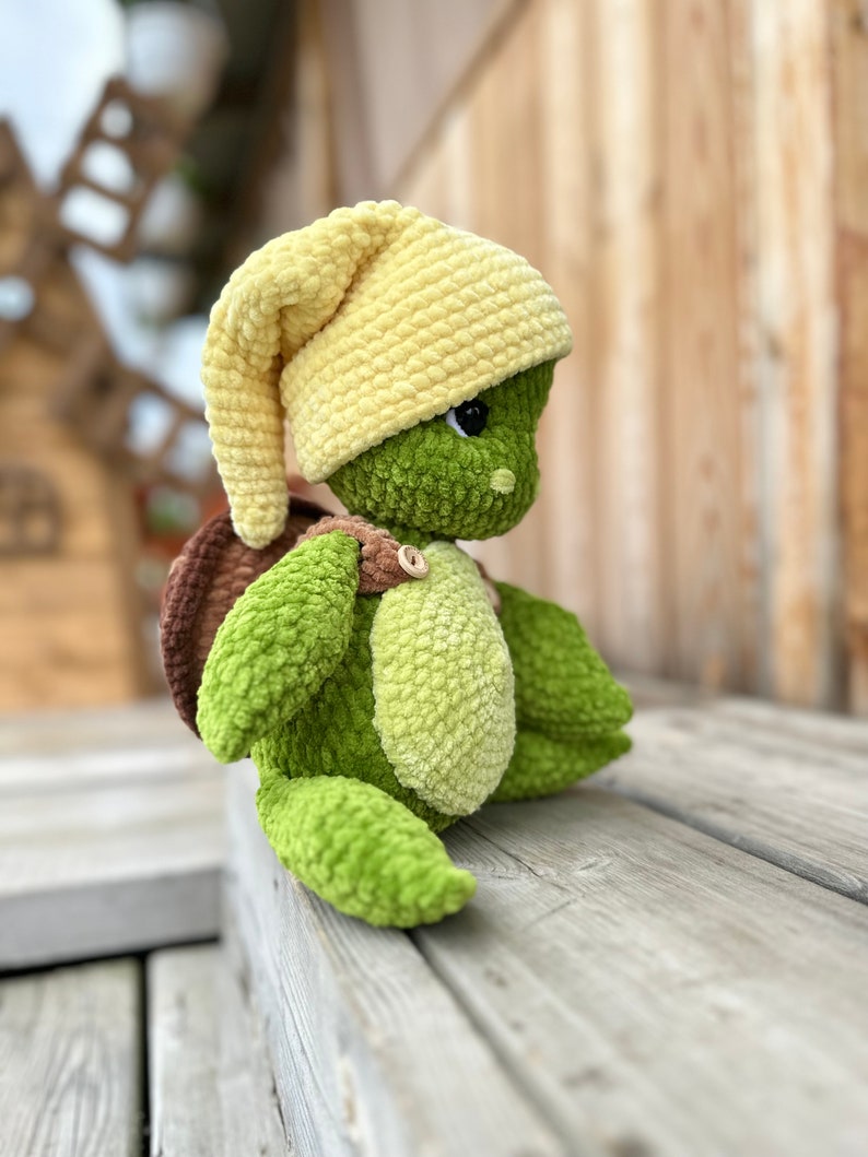 Puede incluir: Un juguete de peluche con forma de tortuga hecho de hilo verde y verde claro, con un caparaz&oacute;n marr&oacute;n y un gorro amarillo suave. La tortuga tiene una cabeza redonda con ojos negros, una mancha verde claro en el vientre y un bot&oacute;n de madera.