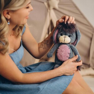 Pu&ograve; includere: Una donna con un vestito di jeans blu tiene in mano un peluche grigio lavorato a uncinetto con dettagli rosa e una coda pelosa. L'animale ha un viso cucito con occhi neri e un naso giallo.