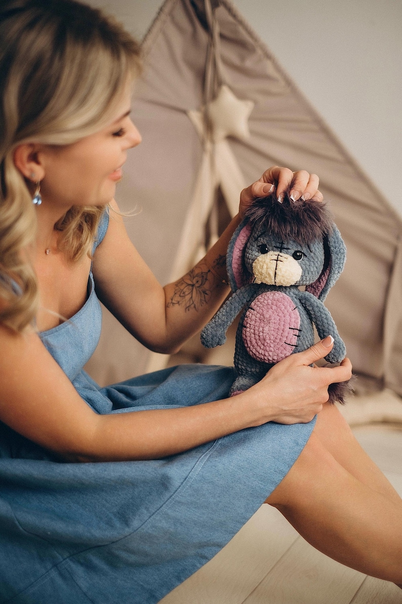 Pu&ograve; includere: Una donna con un vestito di jeans blu tiene in mano un peluche grigio lavorato a uncinetto con dettagli rosa e una coda pelosa. L'animale ha un viso cucito con occhi neri e un naso giallo.