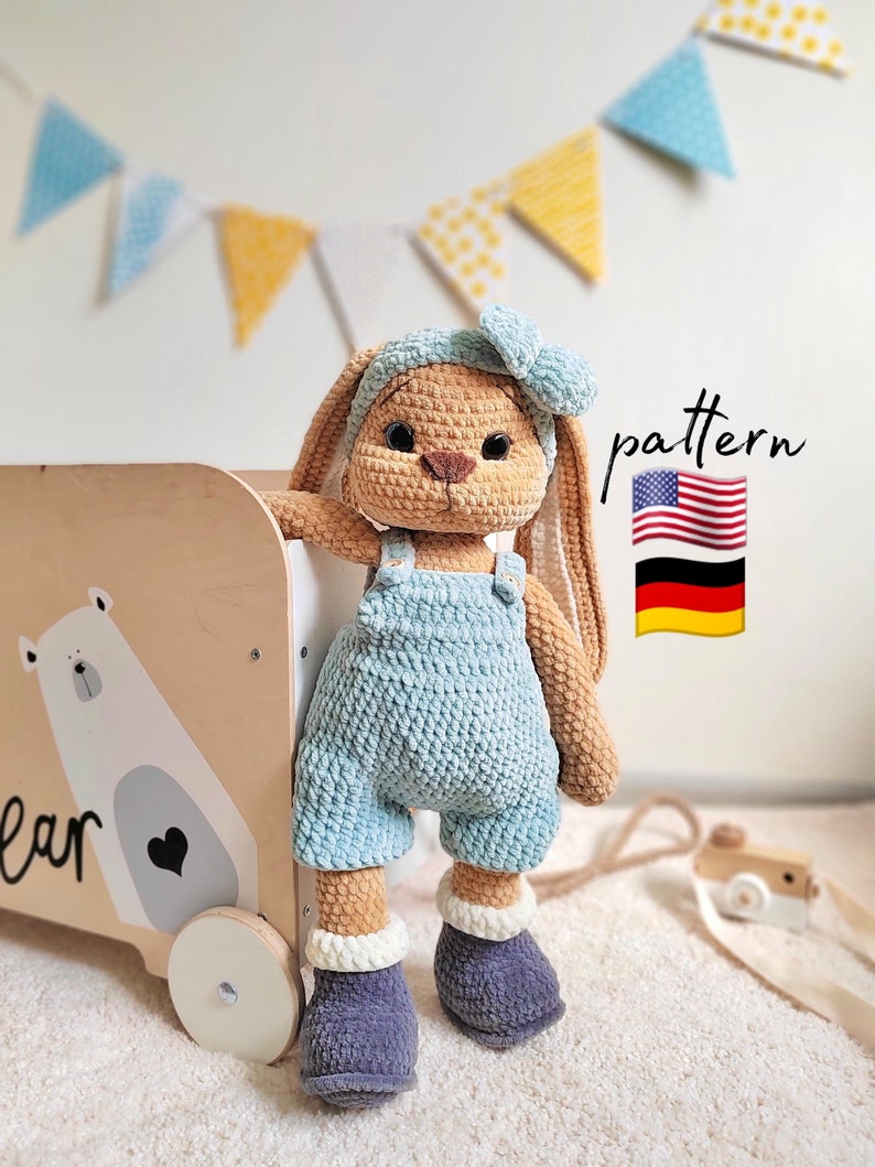 May include: A crocheted tan rabbit plush toy wearing light blue overalls, a matching headband with a bow, and dark gray boots. The rabbit stands next to a wooden toy cart featuring a bear illustration and the text pattern with US and German flags.