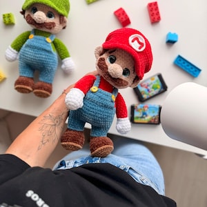 May include: Two handmade crochet plush dolls representing Mario and Luigi. Each figure features a signature colored cap with a white letter emblem, a brown mustache, blue overalls with yellow buttons, white gloves, and brown shoes. The dolls are crafted with soft yarn.