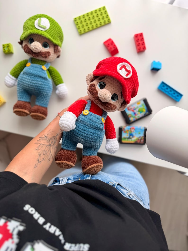 May include: Two handmade crochet plush dolls representing Mario and Luigi. Each figure features a signature colored cap with a white letter emblem, a brown mustache, blue overalls with yellow buttons, white gloves, and brown shoes. The dolls are crafted with soft yarn.