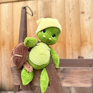 Puede incluir: Un juguete de peluche con forma de tortuga hecho de hilo verde y verde claro, con un caparaz&oacute;n marr&oacute;n, un gorro amarillo y dos peque&ntilde;os botones de madera en el pecho. La tortuga tiene grandes ojos negros y una textura suave y visible.