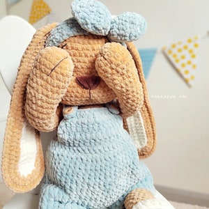 May include: A plush crochet bunny toy with tan fur, wearing light blue overalls and a matching headband. The bunny has long ears with white inner sections and is covering its eyes with its paws. The text PARKAEVA_HM is visible on the side.