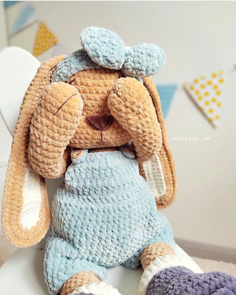 May include: A plush crochet bunny toy with tan fur, wearing light blue overalls and a matching headband. The bunny has long ears with white inner sections and is covering its eyes with its paws. The text PARKAEVA_HM is visible on the side.