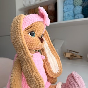 May include: A plush crochet rabbit doll with tan fur, long ears, and a pink headband. The doll wears a pink romper with small buttons and pink boots with white cuffs. It features large blue eyes with long eyelashes and a dark brown nose.