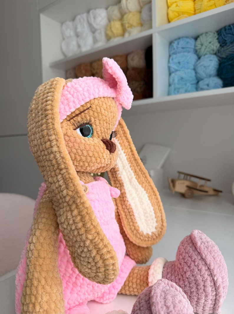 May include: A plush crochet rabbit doll with tan fur, long ears, and a pink headband. The doll wears a pink romper with small buttons and pink boots with white cuffs. It features large blue eyes with long eyelashes and a dark brown nose.
