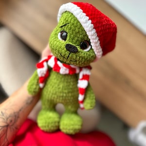 Peut inclure: Une poup&eacute;e en peluche crochet&eacute;e verte avec de grands yeux sombres et un nez noir brod&eacute;. La poup&eacute;e porte un bonnet de P&egrave;re No&euml;l crochet&eacute; rouge et blanc et une &eacute;charpe assortie ray&eacute;e rouge et blanche autour du cou, faite de fil textur&eacute; doux.