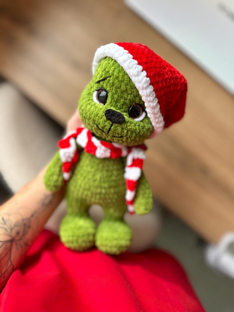 Peut inclure: Une poup&eacute;e en peluche crochet&eacute;e verte avec de grands yeux sombres et un nez noir brod&eacute;. La poup&eacute;e porte un bonnet de P&egrave;re No&euml;l crochet&eacute; rouge et blanc et une &eacute;charpe assortie ray&eacute;e rouge et blanche autour du cou, faite de fil textur&eacute; doux.