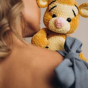 May include: A plush stuffed animal resembling a tiger, crafted from golden-yellow and cream-coloured yarn with a soft, textured finish. The toy features black embroidered eyebrows, small black eyes, and a light pink nose, with rounded ears on top.