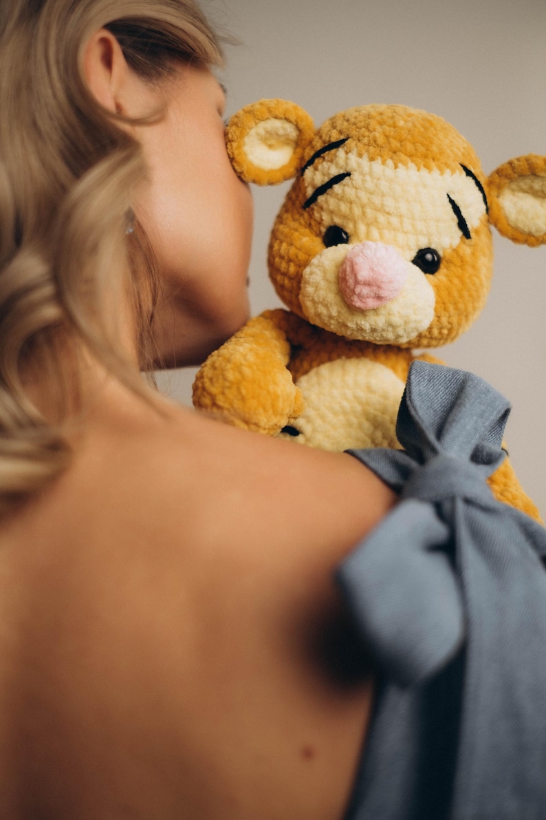 May include: A plush stuffed animal resembling a tiger, crafted from golden-yellow and cream-coloured yarn with a soft, textured finish. The toy features black embroidered eyebrows, small black eyes, and a light pink nose, with rounded ears on top.