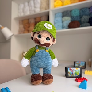 May include: A plush crochet doll of a character wearing a green cap with a white L emblem, a green long-sleeve shirt, blue overalls with yellow buttons, white gloves, and brown shoes. The doll has large blue eyes and a brown mustache.