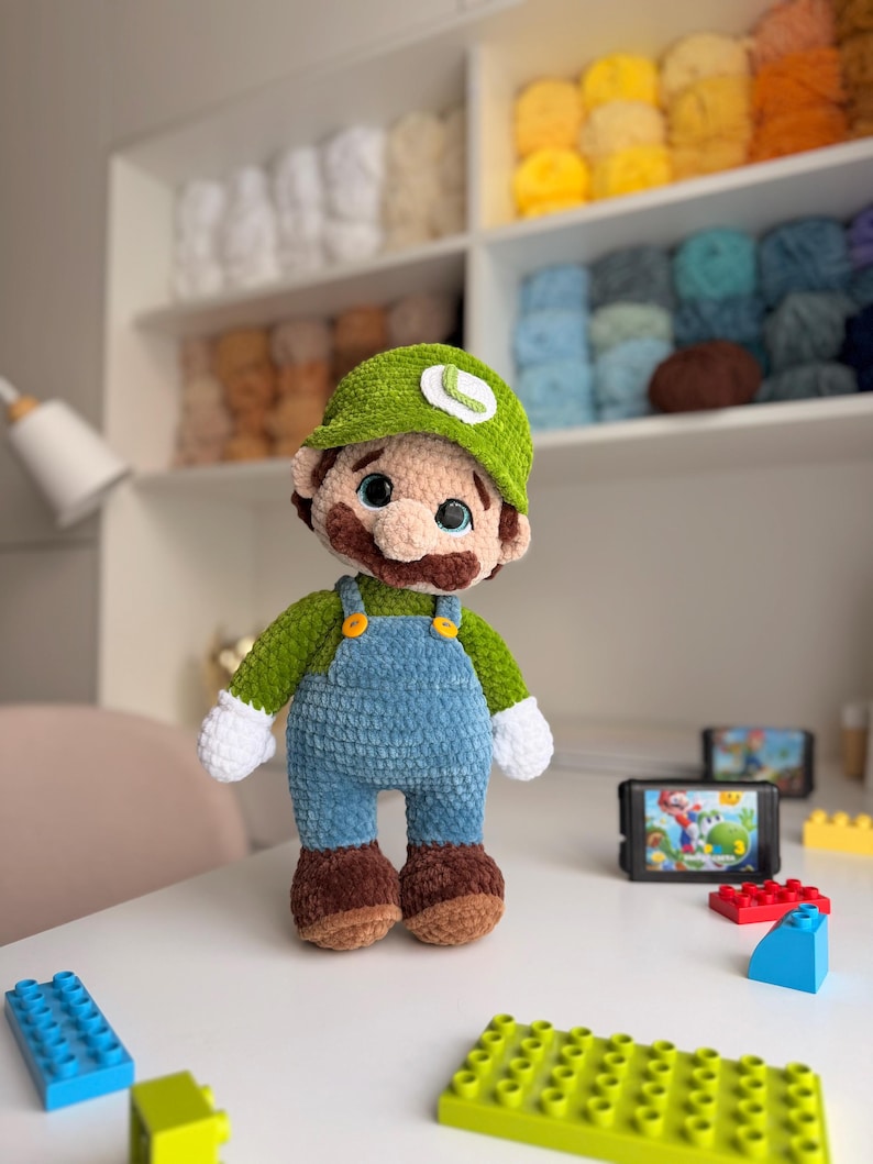 May include: A plush crochet doll of a character wearing a green cap with a white L emblem, a green long-sleeve shirt, blue overalls with yellow buttons, white gloves, and brown shoes. The doll has large blue eyes and a brown mustache.