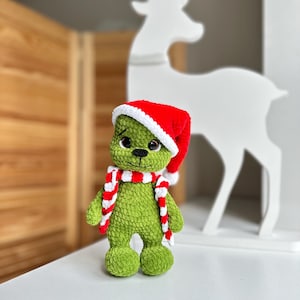 Peut inclure: Une peluche crochet&eacute;e verte &agrave; la texture douce et duveteuse, dot&eacute;e de grands yeux sombres et d'un petit nez noir. La figurine porte un bonnet de P&egrave;re No&euml;l rouge vif avec une bordure blanche et une &eacute;charpe ray&eacute;e rouge et blanche assortie.