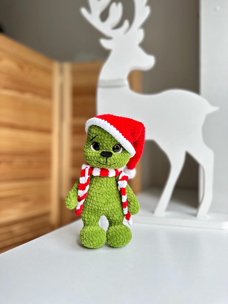 Peut inclure: Une peluche crochet&eacute;e verte &agrave; la texture douce et duveteuse, dot&eacute;e de grands yeux sombres et d'un petit nez noir. La figurine porte un bonnet de P&egrave;re No&euml;l rouge vif avec une bordure blanche et une &eacute;charpe ray&eacute;e rouge et blanche assortie.