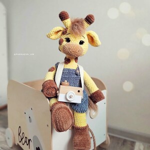 May include: A crocheted giraffe toy wearing overalls and holding a toy camera. The giraffe is sitting on a wooden toy box with the word "bear" written on it.