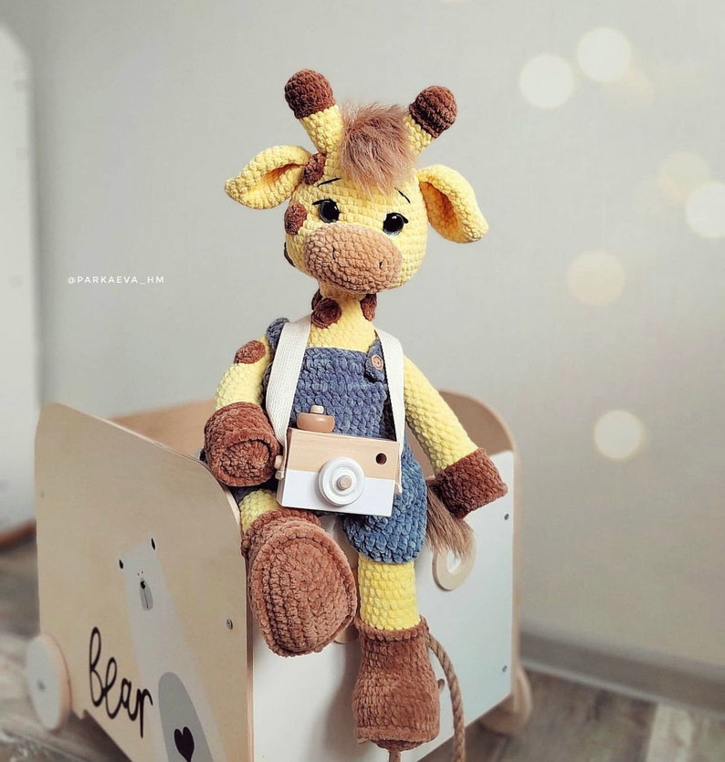 May include: A crocheted giraffe toy wearing overalls and holding a toy camera. The giraffe is sitting on a wooden toy box with the word "bear" written on it.