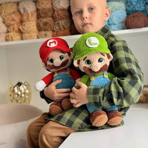May include: Two handmade crochet plush dolls representing Mario and Luigi. Mario wears a red hat with a white M, a red shirt, and blue overalls. Luigi wears a green hat with a white L, a green shirt, and blue overalls. Both have brown mustaches.