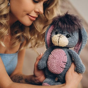 Pu&ograve; includere: Una donna tiene tra le braccia un peluche all'uncinetto. L'animale &egrave; grigio con dettagli rosa e ha una lunga coda pelosa. La donna indossa un vestito blu e ha lunghi capelli biondi.