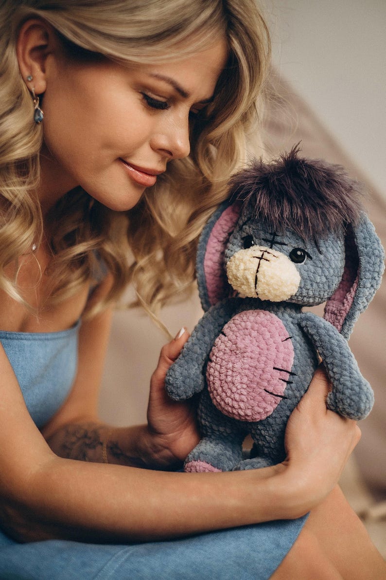 Pu&ograve; includere: Una donna tiene tra le braccia un peluche all'uncinetto. L'animale &egrave; grigio con dettagli rosa e ha una lunga coda pelosa. La donna indossa un vestito blu e ha lunghi capelli biondi.