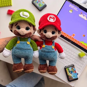 May include: Two plush crochet dolls representing video game characters. One doll wears a green hat with an L and green shirt, the other a red hat with an M and red shirt. Both have blue overalls with yellow buttons, brown shoes, and white gloves.