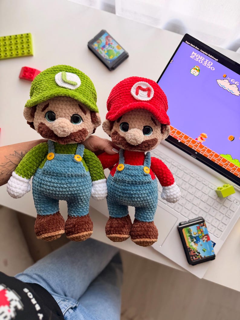 May include: Two plush crochet dolls representing video game characters. One doll wears a green hat with an L and green shirt, the other a red hat with an M and red shirt. Both have blue overalls with yellow buttons, brown shoes, and white gloves.