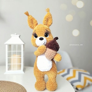 May include: A crocheted squirrel plush toy with large eyes and a brown acorn in its paws. The squirrel is brown and white and has a fluffy tail.