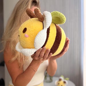 K&ouml;nnte beinhalten: Ein gro&szlig;es, pl&uuml;schiges Bienenspielzeug. Es hat gelbe und braune Streifen, wei&szlig;e Fl&uuml;gel und ein gr&uuml;nes Blatt. Die Biene hat ein l&auml;chelndes Gesicht und braune F&uuml;hler. Das Spielzeug ist weich und rund.