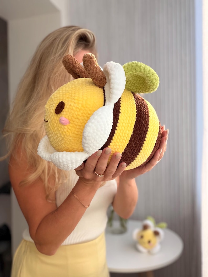 K&ouml;nnte beinhalten: Ein gro&szlig;es, pl&uuml;schiges Bienenspielzeug. Es hat gelbe und braune Streifen, wei&szlig;e Fl&uuml;gel und ein gr&uuml;nes Blatt. Die Biene hat ein l&auml;chelndes Gesicht und braune F&uuml;hler. Das Spielzeug ist weich und rund.