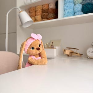May include: A tan crocheted bunny doll with long ears, wearing a pink headband with a bow and a pink outfit. The doll has large dark eyes, a brown nose, and is positioned with its arms resting on a white surface, appearing to look forward.