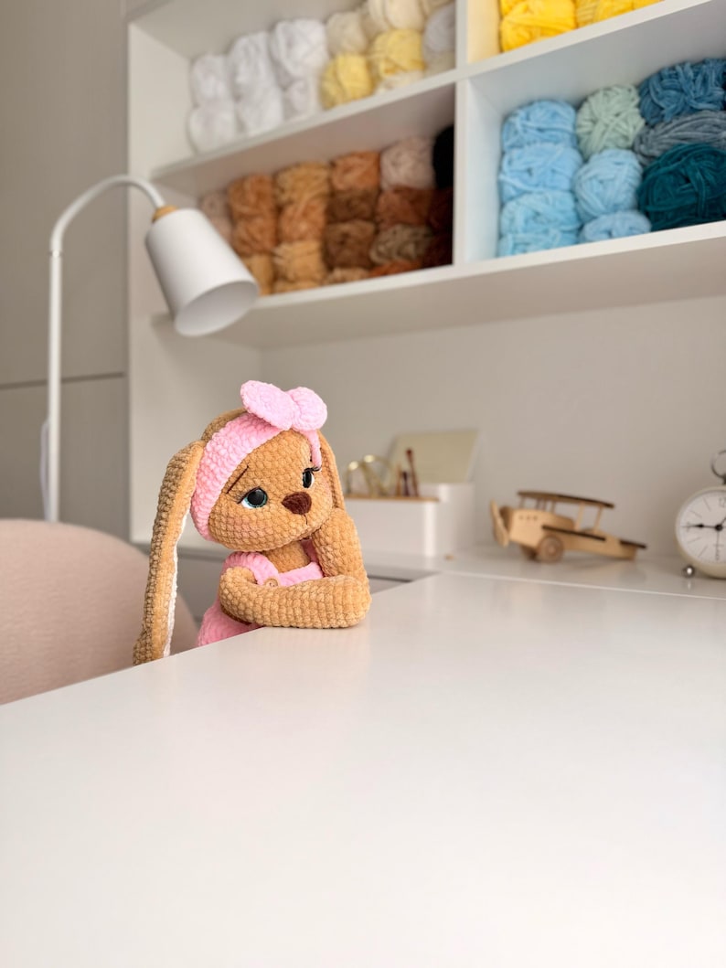 May include: A tan crocheted bunny doll with long ears, wearing a pink headband with a bow and a pink outfit. The doll has large dark eyes, a brown nose, and is positioned with its arms resting on a white surface, appearing to look forward.