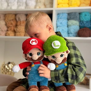 May include: Two handmade crochet plush dolls representing Mario and Luigi. Mario wears a red hat with an M, a red shirt, and blue overalls. Luigi wears a green hat with an L, a green shirt, and blue overalls. Both have brown mustaches.