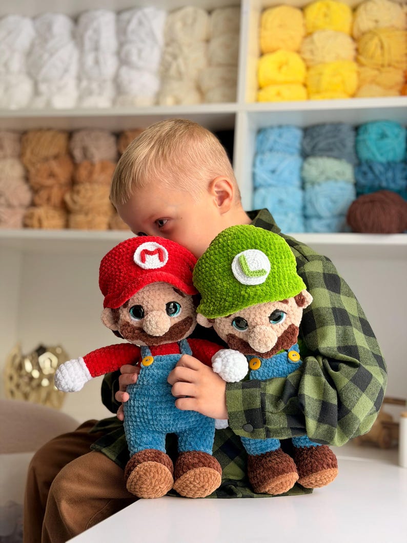 May include: Two handmade crochet plush dolls representing Mario and Luigi. Mario wears a red hat with an M, a red shirt, and blue overalls. Luigi wears a green hat with an L, a green shirt, and blue overalls. Both have brown mustaches.