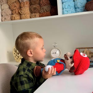 May include: A handcrafted crochet doll of a character wearing a red hat with a white emblem, blue overalls with yellow buttons, and brown shoes. The doll features a tan face, brown mustache, and white gloves, constructed from soft yarn.