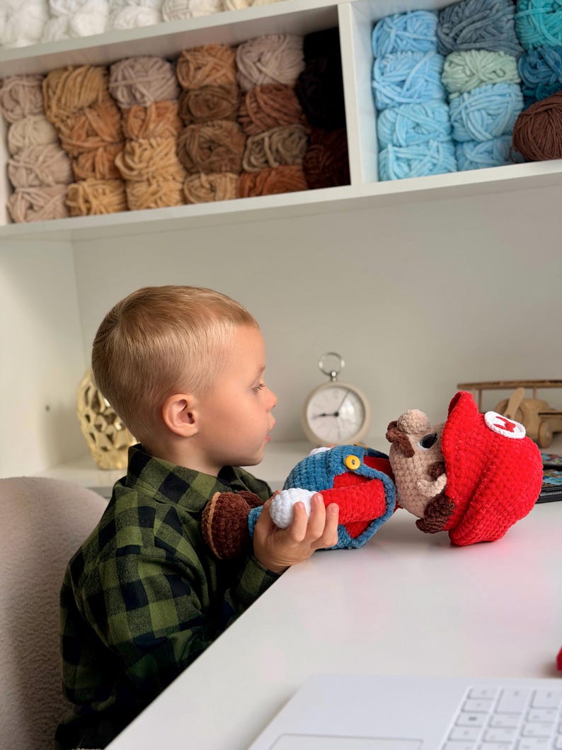 May include: A handcrafted crochet doll of a character wearing a red hat with a white emblem, blue overalls with yellow buttons, and brown shoes. The doll features a tan face, brown mustache, and white gloves, constructed from soft yarn.