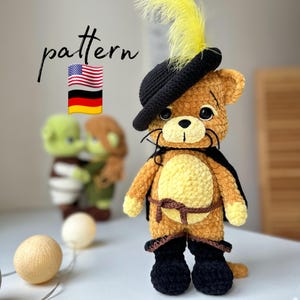 May include: Crocheted cat toy wearing a black hat with a yellow feather, brown pants, and black boots. The cat is holding a brown sword and has a yellow cape. The toy is standing on a white surface. The word "pattern" is written above the toy in black letters.