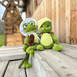 Puede incluir: Dos peluches de ganchillo hechos a mano, una rana verde con sombrero blanco que sostiene una espada&ntilde;a marr&oacute;n, y una tortuga verde con sombrero amarillo y peto marr&oacute;n con botones peque&ntilde;os, ambos con grandes ojos oscuros y lana texturizada.