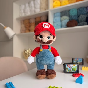 May include: A plush crochet doll of the character Mario, featuring a red hat with a white M emblem, a red shirt, blue overalls with yellow buttons, white gloves, and brown shoes. The doll has large dark eyes and a brown mustache.