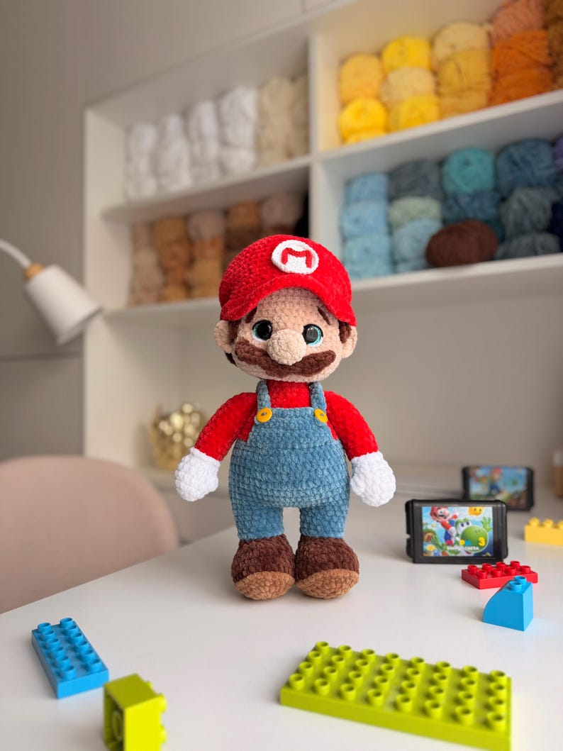 May include: A plush crochet doll of the character Mario, featuring a red hat with a white M emblem, a red shirt, blue overalls with yellow buttons, white gloves, and brown shoes. The doll has large dark eyes and a brown mustache.