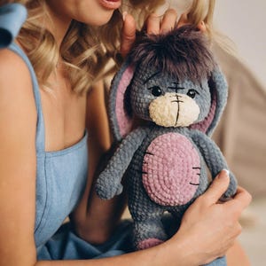 Pu&ograve; includere: Una donna con un vestito blu tiene un peluche grigio e rosa all'uncinetto con grandi orecchie. L'animale ha un viso cucito con occhi neri e una bocca cucita.