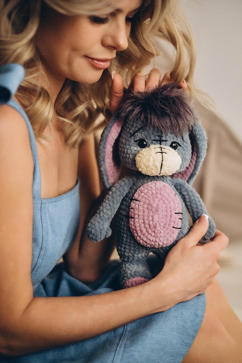 Pu&ograve; includere: Una donna con un vestito blu tiene un peluche grigio e rosa all'uncinetto con grandi orecchie. L'animale ha un viso cucito con occhi neri e una bocca cucita.