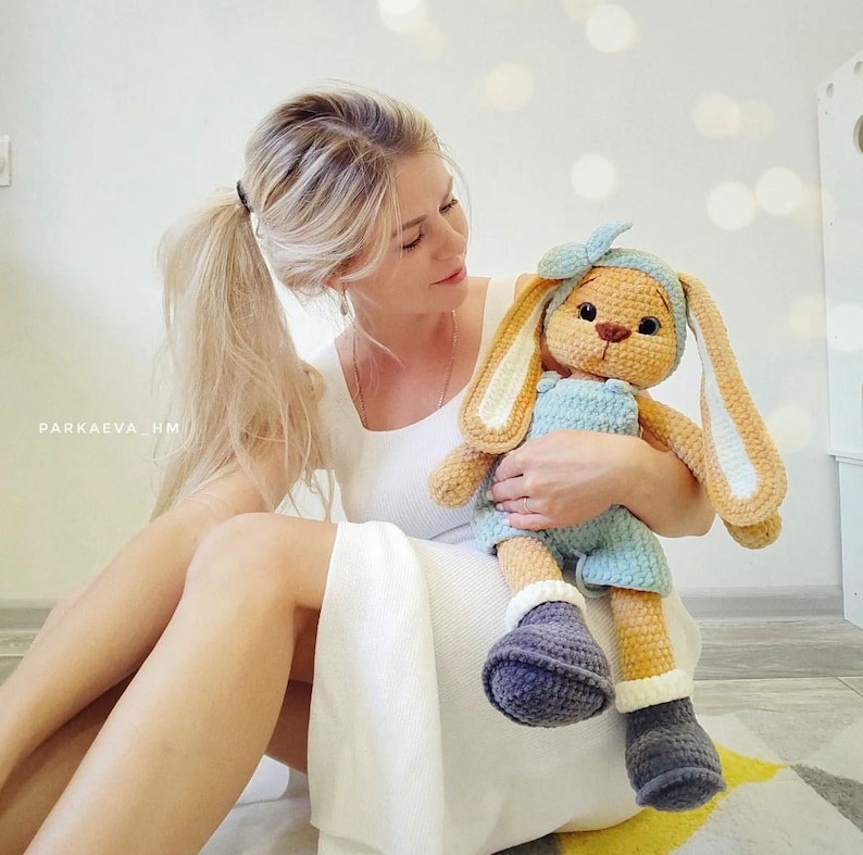 May include: A crocheted stuffed rabbit toy with long ears, wearing a light blue romper, a matching headband with a bow, and dark gray boots with white cuffs. The rabbit has a tan body, dark eyes, and a brown stitched nose. Text reads PARKAEVA_HM.