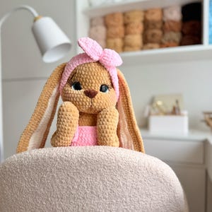 May include: A tan crochet bunny plush toy with long ears, wearing a pink headband with a bow and a pink garment. The bunny has large dark eyes with long eyelashes, a brown nose, and is crafted from soft, textured yarn material.