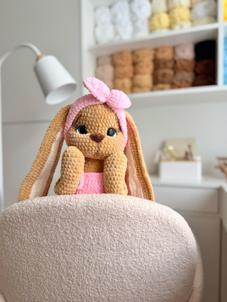 May include: A tan crochet bunny plush toy with long ears, wearing a pink headband with a bow and a pink garment. The bunny has large dark eyes with long eyelashes, a brown nose, and is crafted from soft, textured yarn material.