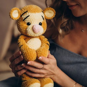 May include: A plush tiger toy made of soft, golden-yellow and cream-coloured yarn. The stuffed animal features black embroidered stripes on its head and body, small round ears, a light pink nose, and dark plastic eyes, with a rounded belly.