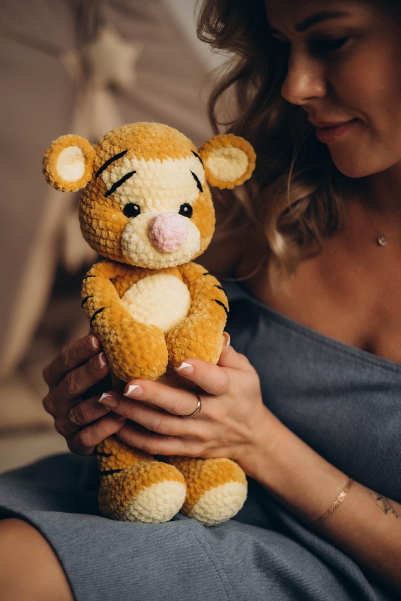 May include: A plush tiger toy made of soft, golden-yellow and cream-coloured yarn. The stuffed animal features black embroidered stripes on its head and body, small round ears, a light pink nose, and dark plastic eyes, with a rounded belly.