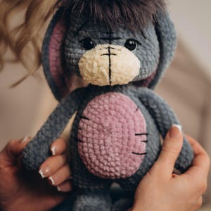 Pu&ograve; includere: Un asino di peluche lavorato a uncinetto con corpo grigio, pancia rosa e pelo nero sulla testa. L'asino ha il naso bianco e gli occhi neri.