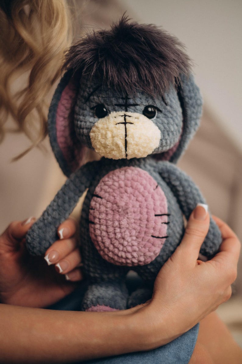Pu&ograve; includere: Un asino di peluche lavorato a uncinetto con corpo grigio, pancia rosa e pelo nero sulla testa. L'asino ha il naso bianco e gli occhi neri.